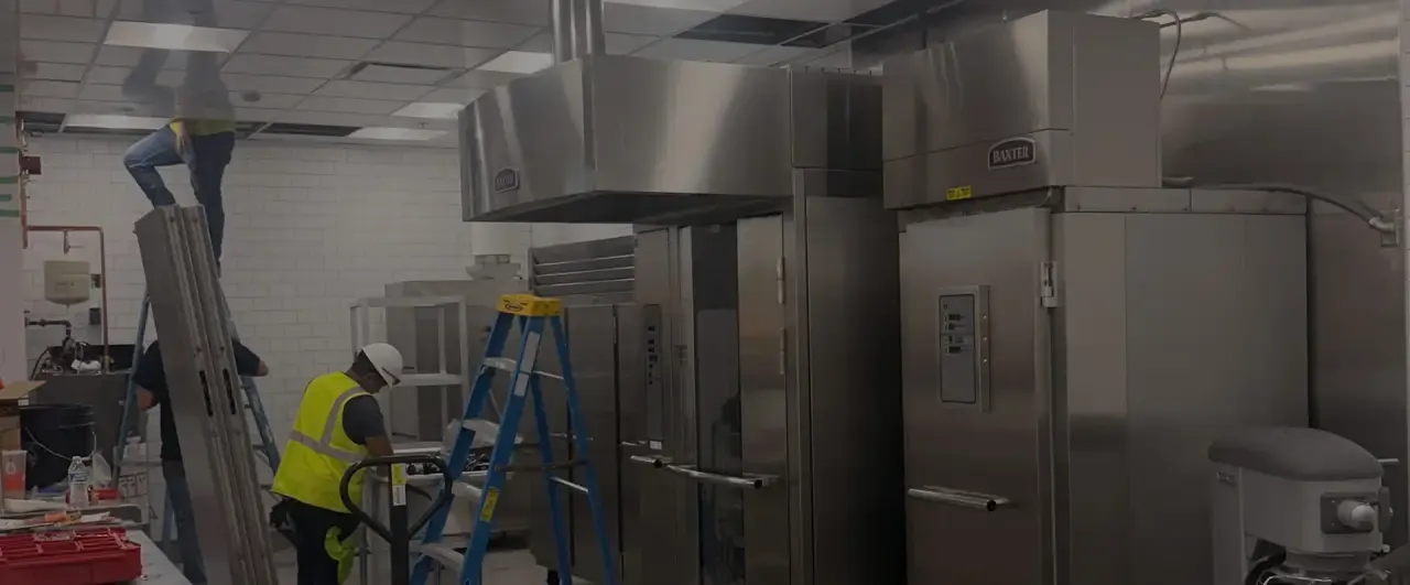 Installer in a hard hat and safety vest working beside new stainless steel rack ovens in a commercial kitchen