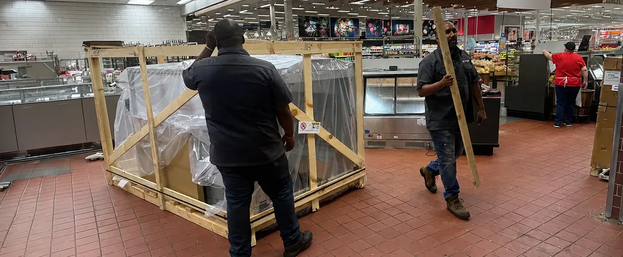 Two installers moving a crated display case across the floor of an open grocery store